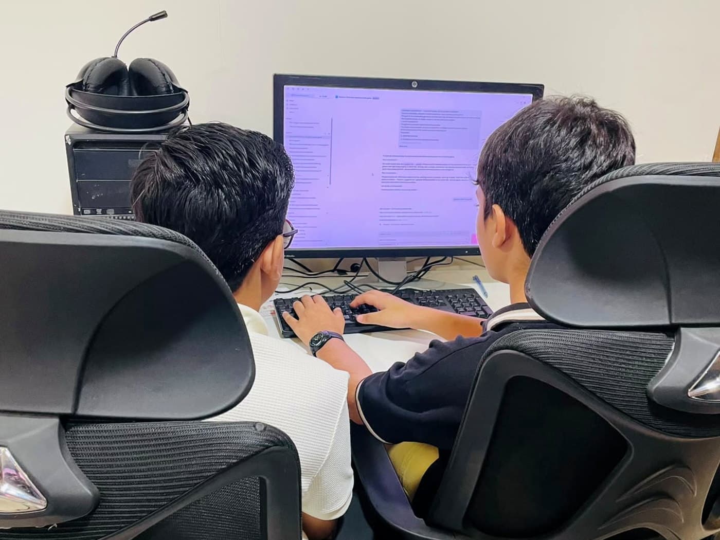 Two children seen from behind at a monitor, one typing, working with an AI assistant on screen