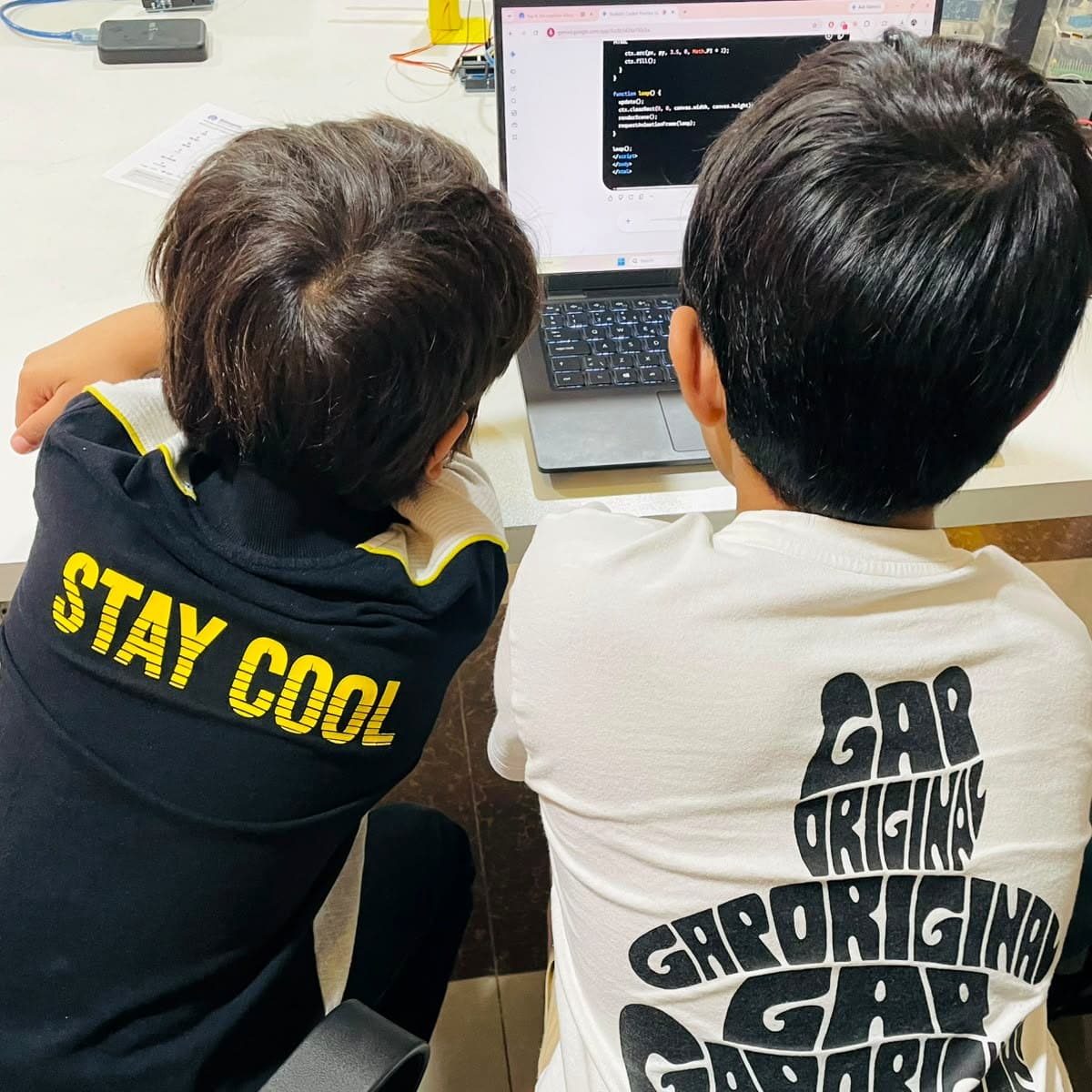 Two children seen from behind leaning over a laptop showing JavaScript canvas code for a browser game