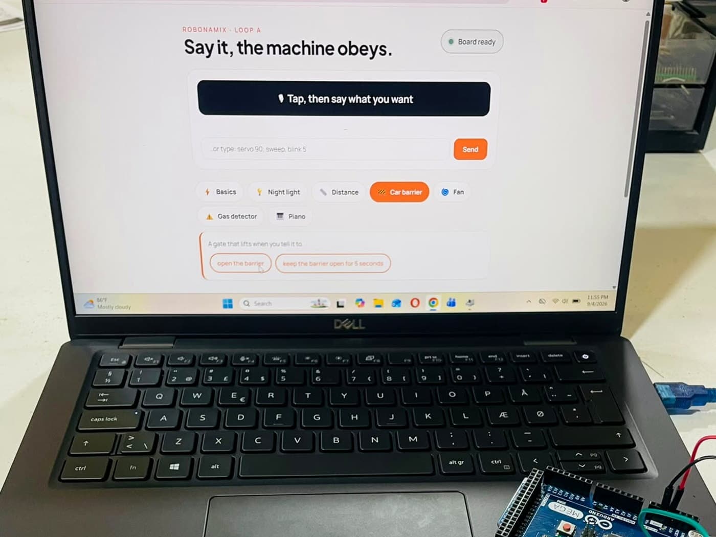 A laptop showing the Robonamix Loop A console reading 'Say it, the machine obeys' with the Car barrier project selected, an Arduino Mega and a cardboard barrier on the desk beside it
