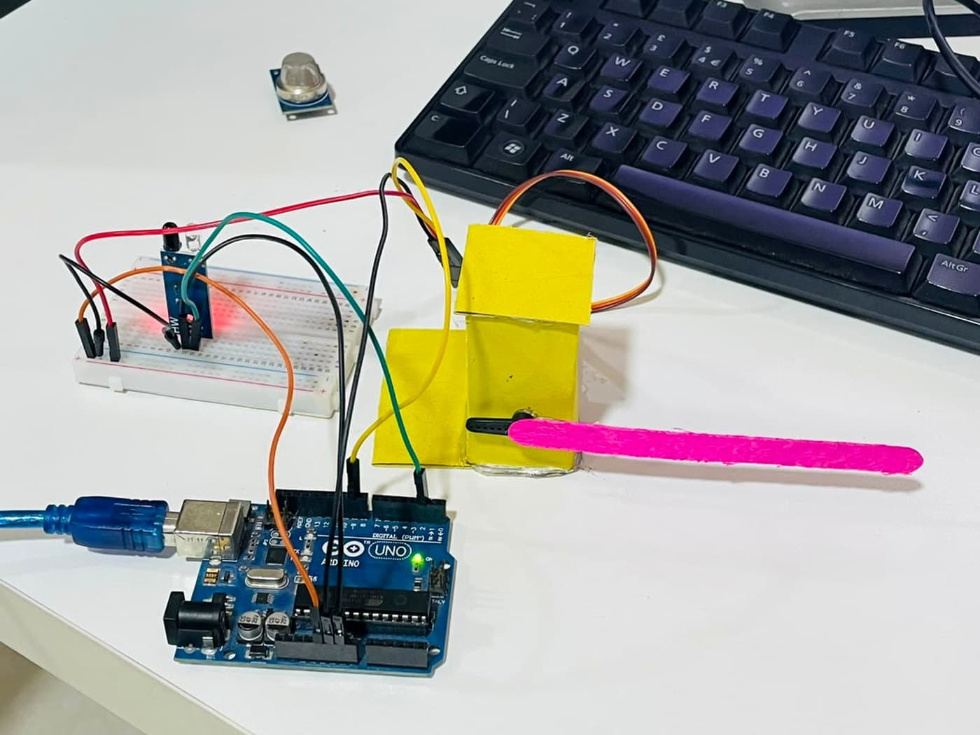 An Arduino Uno wired on a breadboard to a cardboard car barrier with a pink lolly stick for an arm, an infrared sensor and an MQ-2 gas sensor on a white desk
