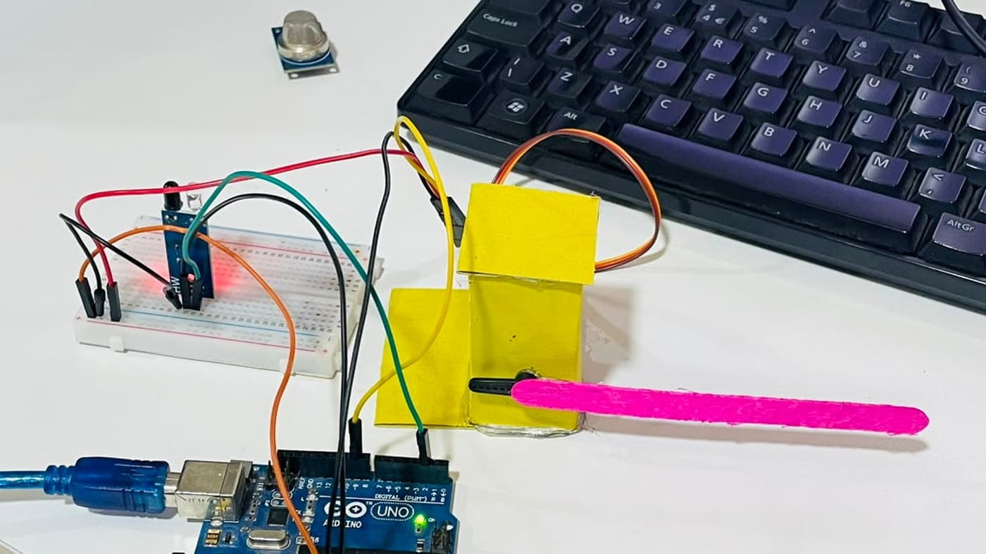 An Arduino Uno wired on a breadboard to a cardboard car barrier with a pink lolly stick for an arm, beside an MQ-2 gas sensor
