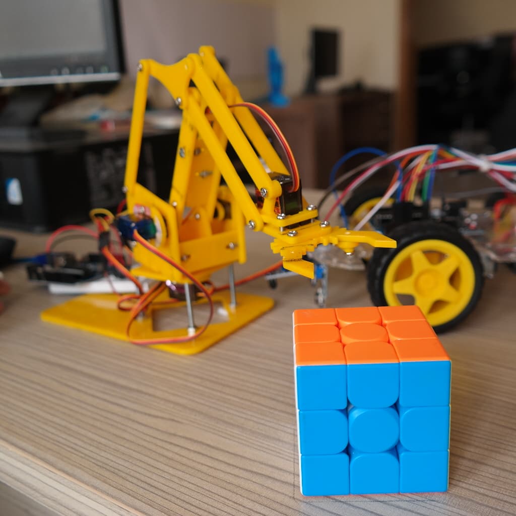 A student-built yellow robot arm, rover and Rubik's cube on the build table